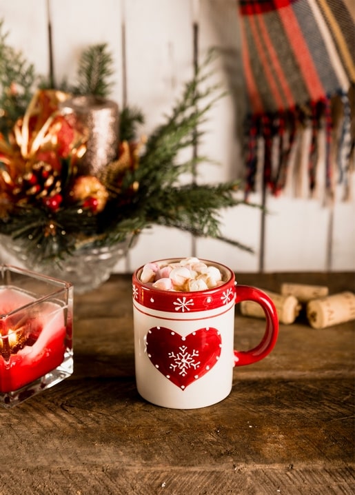 Red heart-patterned mug of cocoa and marshmallows beside a candle and pine arrangement