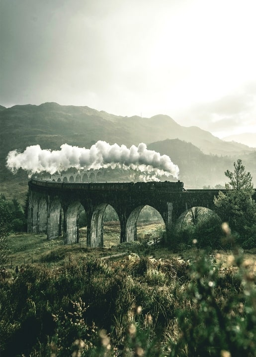 Vintage steam train crossing a stone viaduct with smoke rising over misty Highland mountains