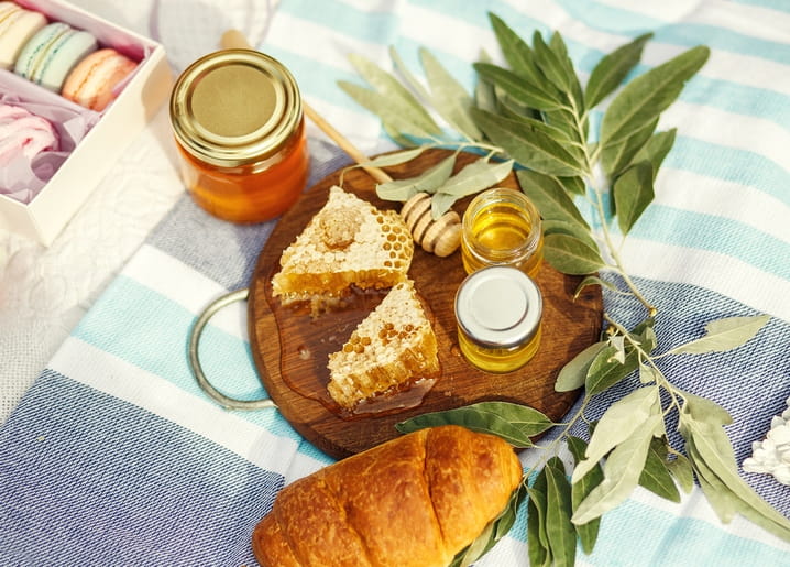 Honigglas, Honigwaben und Croissant auf gestreifter Picknickdecke mit Olivenzweigen