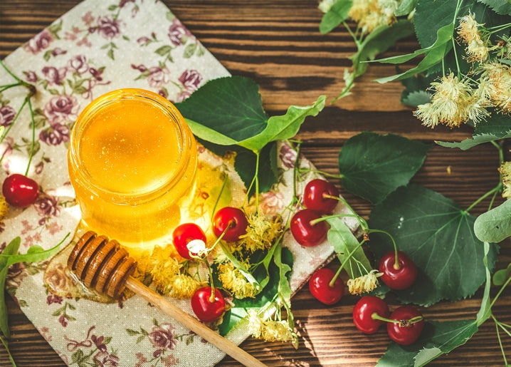 Jar of honey with a wooden dipper, cherries and linden blossoms on a table
