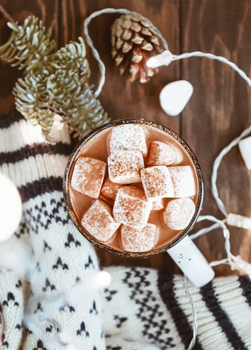 Overhead view of a hot cocoa mug topped with marshmallows next to a pinecone and knit sweater