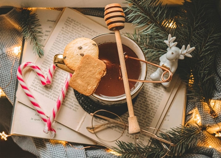 Mug of hot cocoa with honey dipper, cookies, and candy cane on an open storybook among pine branches and fairy lights