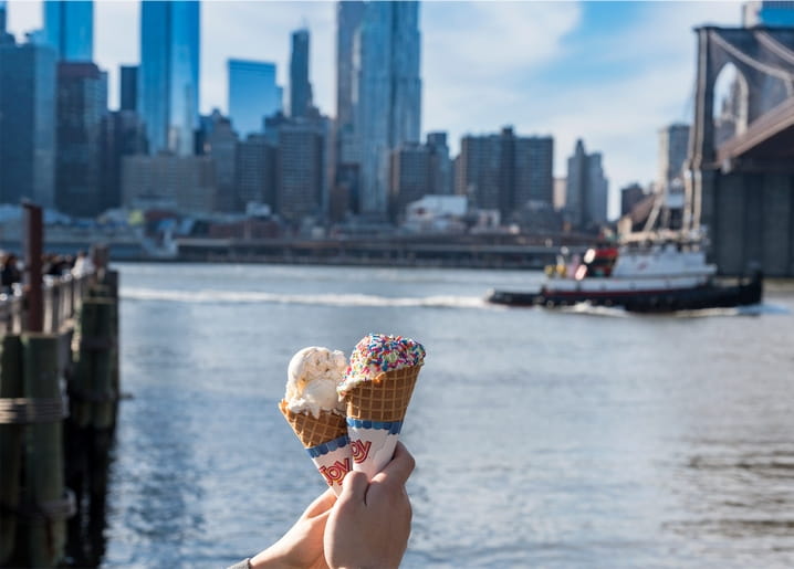 Hand hält zwei Eistüten vor der Brooklyn Bridge und der Manhattan-Skyline