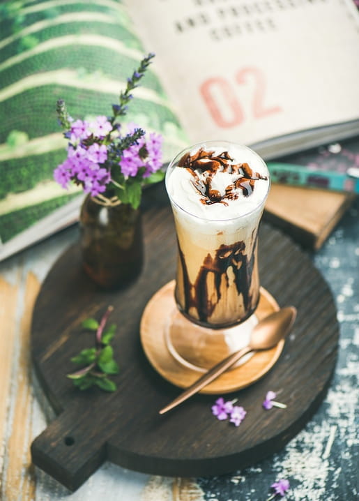 Eiskaffee mit Sahnehaube und Schokosoße neben lila Verbena-Blüten