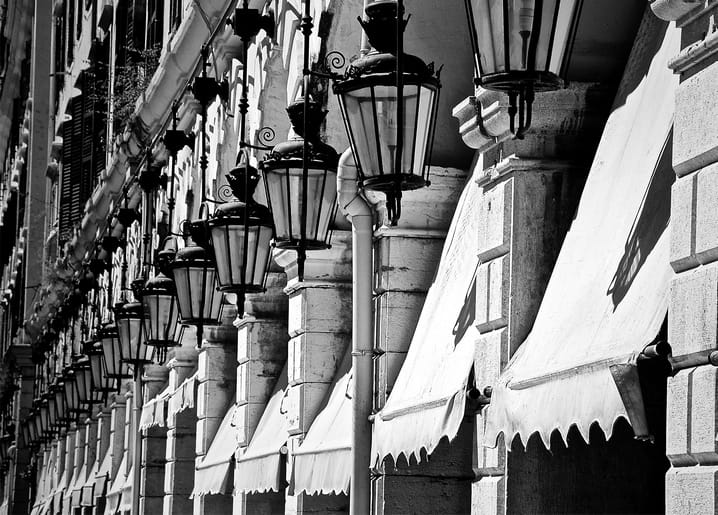 Black-and-white photo of ornate iron lanterns along a sunlit arcade with awnings