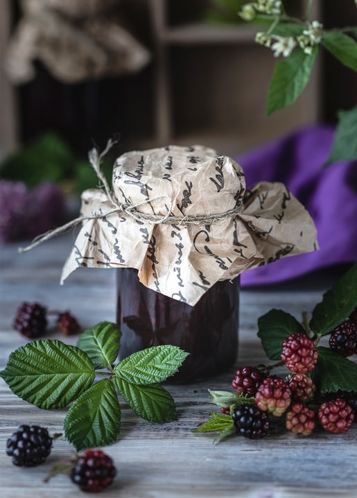 Mit Papier umwickeltes Marmeladenglas, mit Schnur gebunden, umgeben von reifen Brombeeren