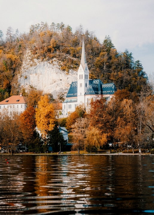 Weiße Kirche mit Turm auf bewaldetem Hügel, gespiegelt in einem stillen See