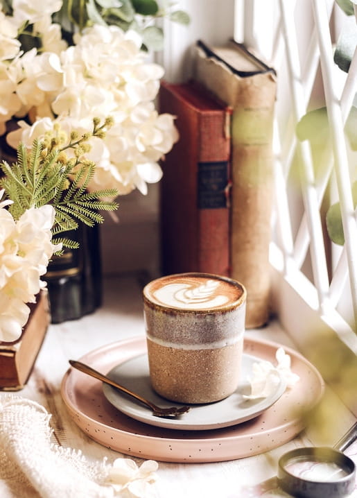 Latte art coffee cup beside vintage books and white blossoms on a windowsill