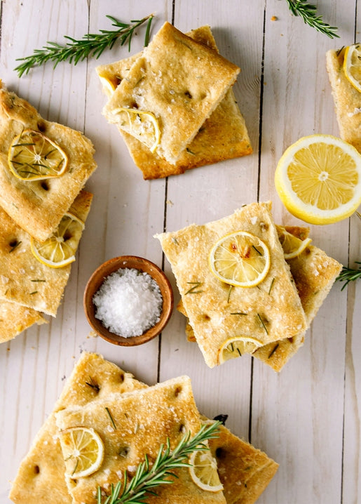 Golden focaccia squares with lemon slices and rosemary sprigs on a wooden board with sea salt.