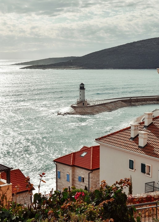 White lighthouse at a harbor entrance below misty mountains and red-tiled rooftops