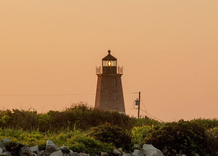 Stone lighthouse lit at dusk above a rocky coastal bluff with wild rose bushes
