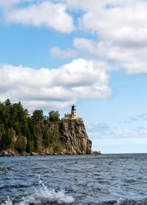 Leuchtturm auf bewaldeter Klippe über blauem Meer mit Wellen und bewölktem Himmel.