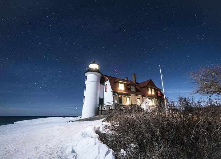 Lit lighthouse beside a shingled house on a snowy coastal bluff under a starry sky