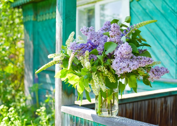 Jar of lilac bouquet on a teal porch railing in front of a cottage window