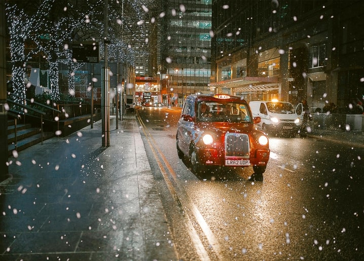 Red London taxi driving through falling snow past illuminated Christmas lights at night