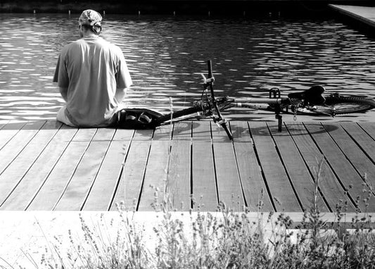 Black-and-white photo of a man sitting on a wooden dock beside his bicycle, facing the water