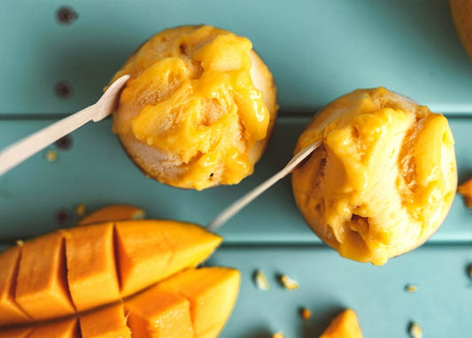 Two scoops of mango ice cream on sticks beside fresh cut mango on a teal table
