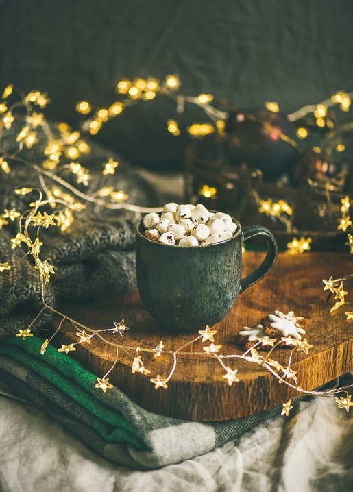 Dark green mug of marshmallows beside star-shaped fairy lights and a grey knit blanket