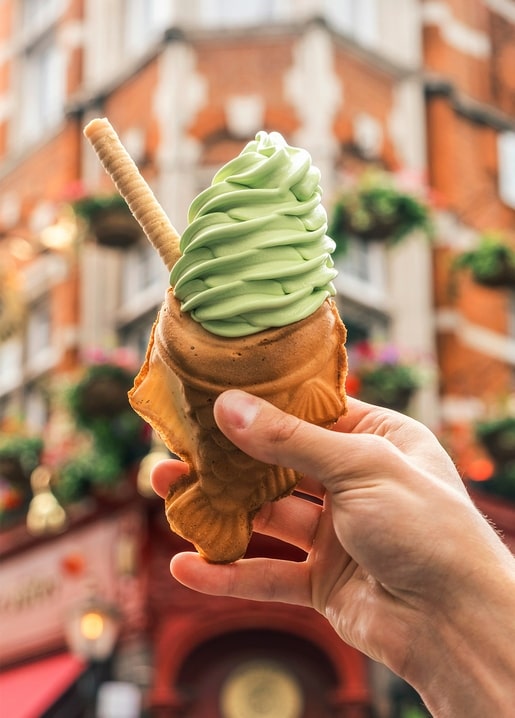 Hand hält fischförmiges Waffelhorn mit grünem Matcha-Softeis auf einer Stadtstraße
