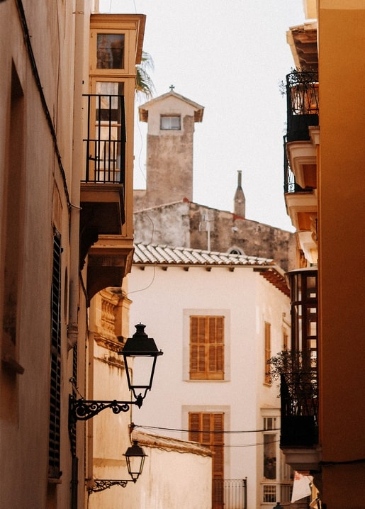 Narrow Mediterranean street with iron balconies, a lantern, and a distant bell tower