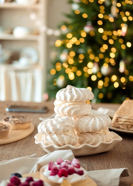 Stacked meringues on a plate with berry tarts and a glowing Christmas tree behind