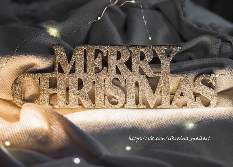 Goldene Glitzerbuchstaben mit dem Schriftzug „Merry Christmas“ auf dunklem Stoff mit Lichterkette