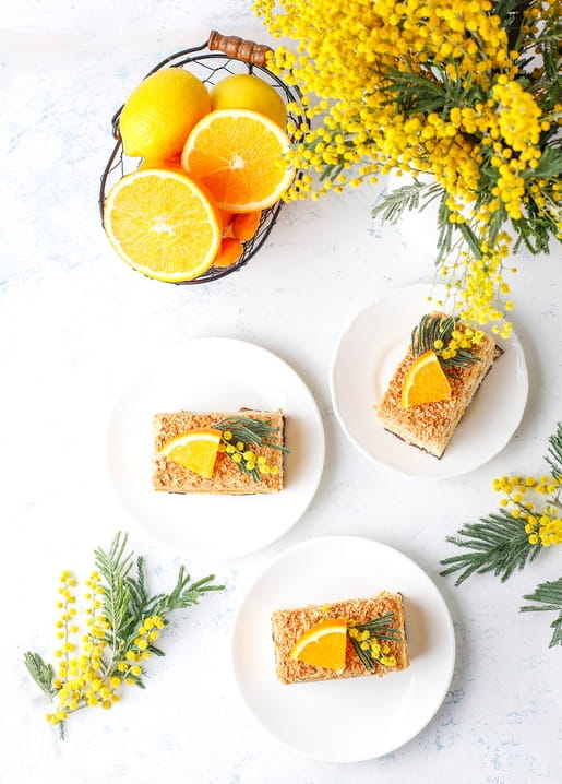 Basket of lemons and oranges beside blooming mimosa, with citrus cake slices on plates