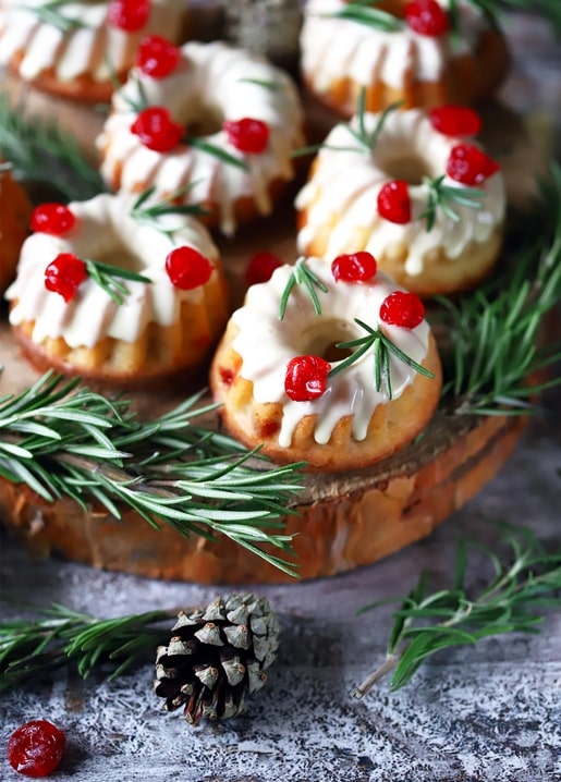 Mini bundt cakes with white icing, cranberries, and rosemary sprigs beside a pinecone