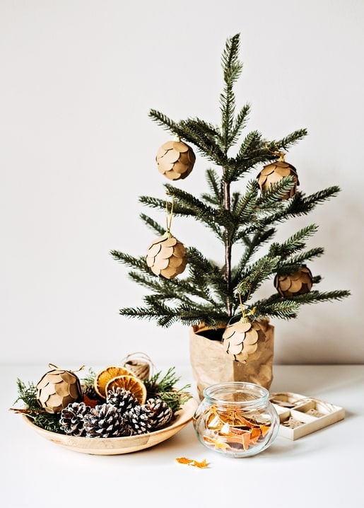 Small potted Christmas tree with paper ornaments beside pinecones and dried orange slices