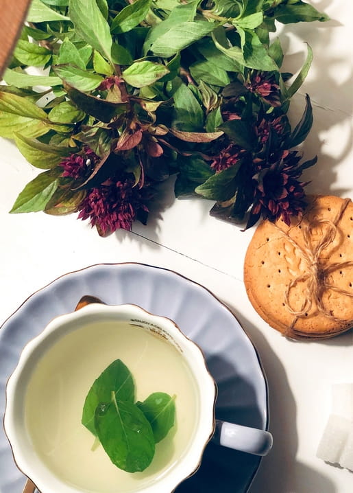 Mint tea in a teacup beside a bouquet of flowering herbs and cookies tied with twine.