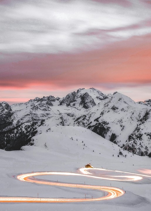 Kurvige Bergstraße mit leuchtenden Lichtspuren unter verschneiten Gipfeln in der Abenddämmerung