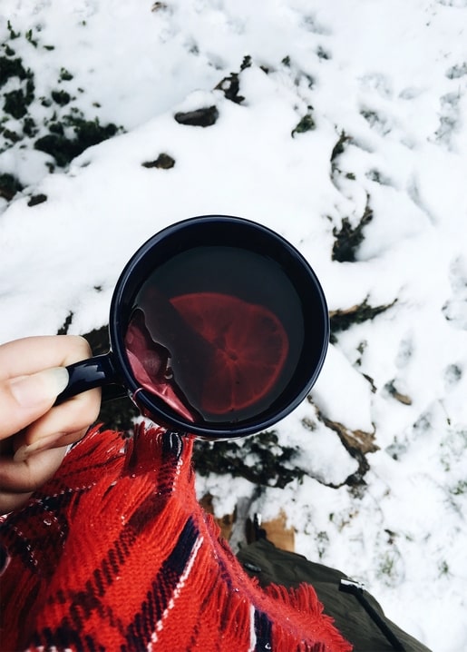 Hand in a red plaid scarf holding a mug of mulled wine with orange slice over snow
