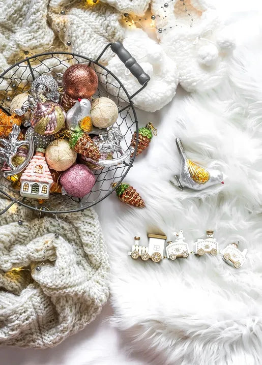Vintage-style Christmas baubles, decorative figurines and pine cones in a black wire basket beside a cream knitted blanket and white faux fur