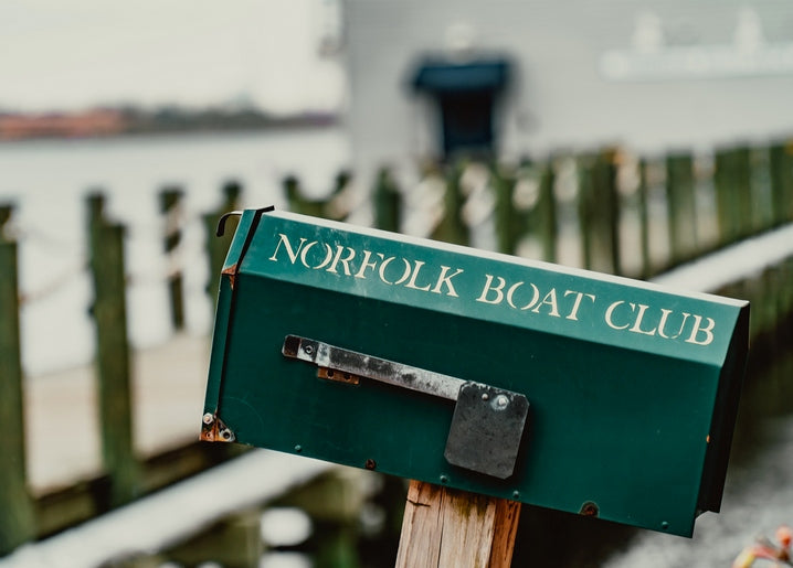 Weathered green mailbox reading Norfolk Boat Club on a wooden dock, boats blurred behind