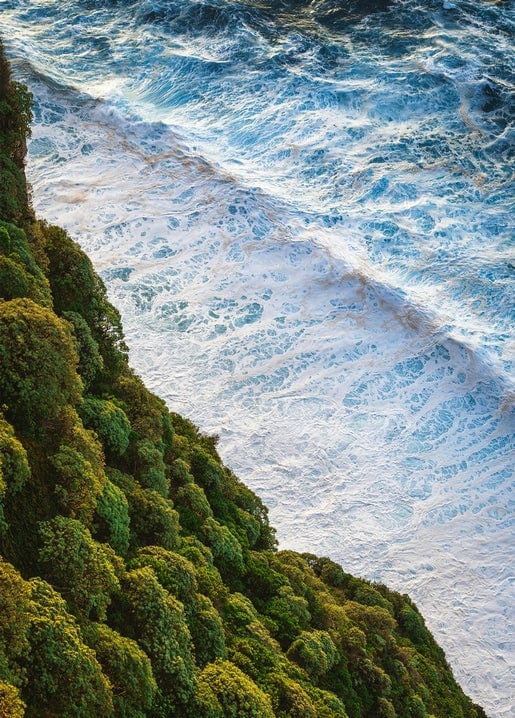 Aerial view of turquoise waves crashing against a forested coastal cliff