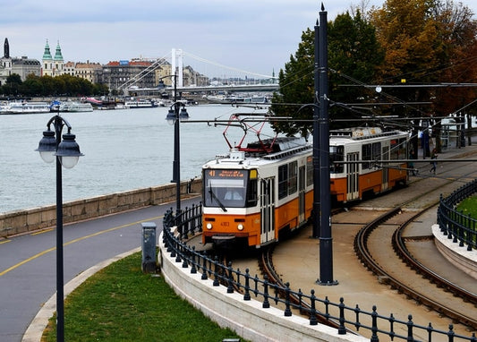 Orangene Straßenbahn an einer Uferpromenade im Herbst, im Hintergrund Brücke und Skyline