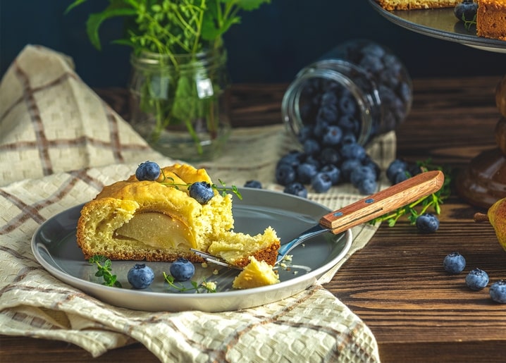 Slice of pear cake with fresh blueberries and thyme sprig on a plate, spilled blueberry jar behind