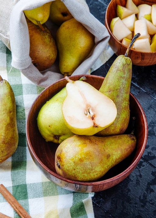 Halved pear beside whole pears in a wooden bowl with cinnamon sticks