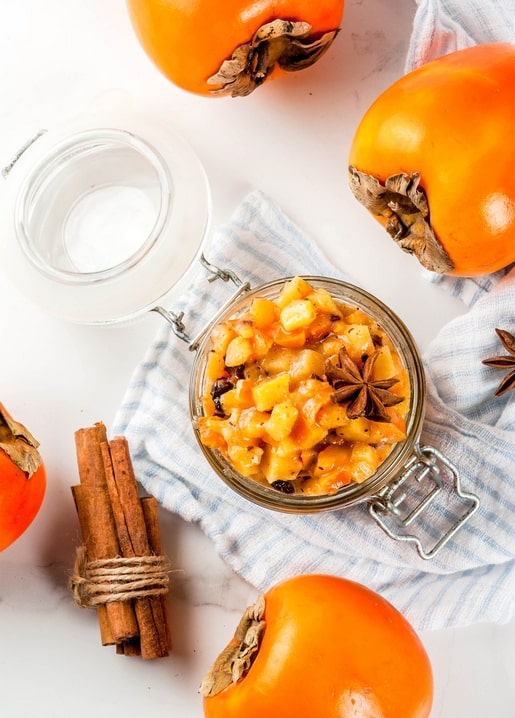 Open jar of persimmon compote with cinnamon sticks and star anise beside whole persimmons