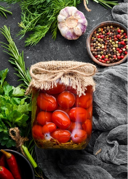 Glas eingelegter Kirschtomaten mit Jute verschnürt, umgeben von Kräutern, Knoblauch und Pfeffer