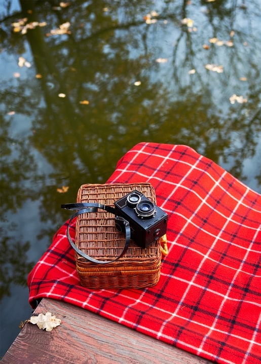 Wicker picnic basket and vintage twin-lens camera on a red plaid blanket by a pond