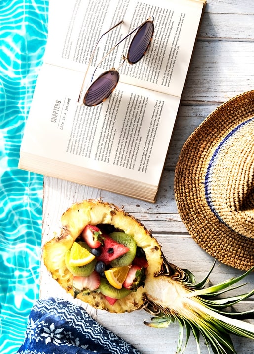 Hollowed pineapple filled with fruit salad beside a book, sunglasses and straw hat by a pool