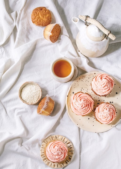 Rosa Cupcakes mit Kokos neben Tee und Muffins auf weißem Leinen