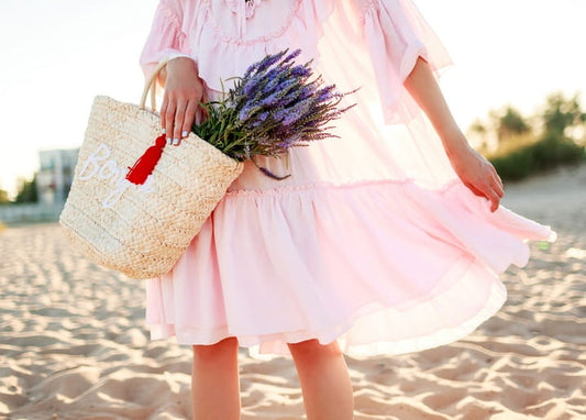 Woman in a pink ruffled dress holding a straw bag and lavender bouquet on a sandy beach