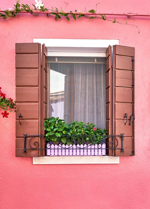 Pink wall with an open window, brown shutters, and a flower box on the sill
