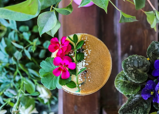 Overhead view of a golden cocktail topped with pink geranium blossoms and greenery on a wooden table