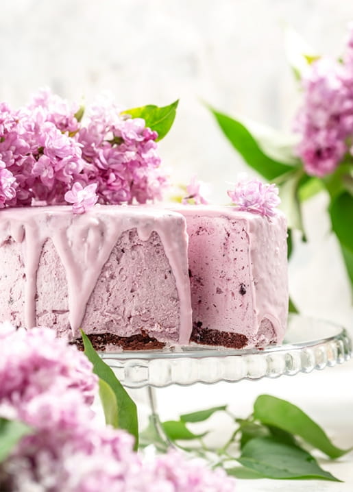 Pink-glazed cake with dripping icing topped with fresh lilac blossoms on a glass cake stand
