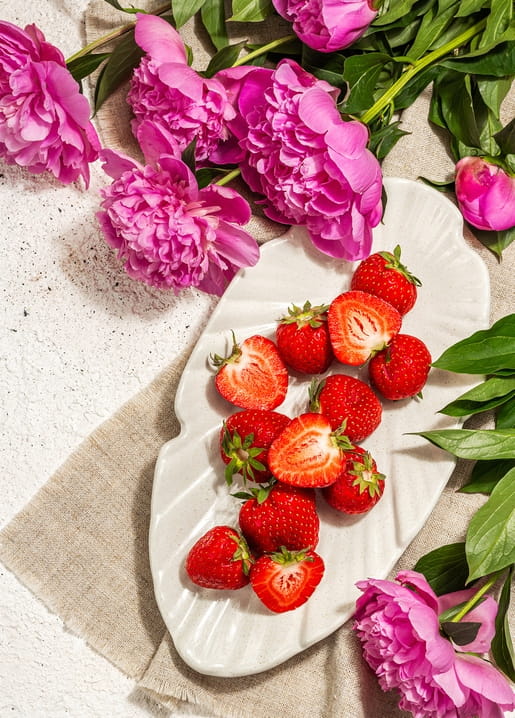 Pink peonies beside a plate of halved strawberries on a linen cloth