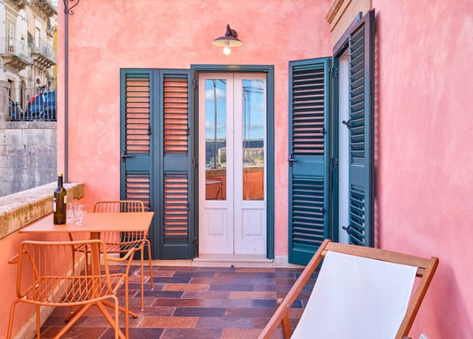 Rosa Terrasse mit türkisen Fensterläden, Weinflasche und Korbsessel