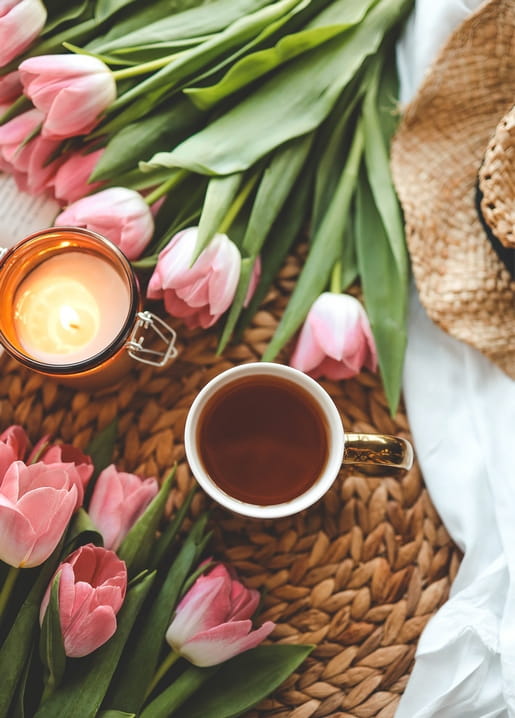 Pink tulips beside a cup of tea, a lit candle, and a straw hat on a woven mat
